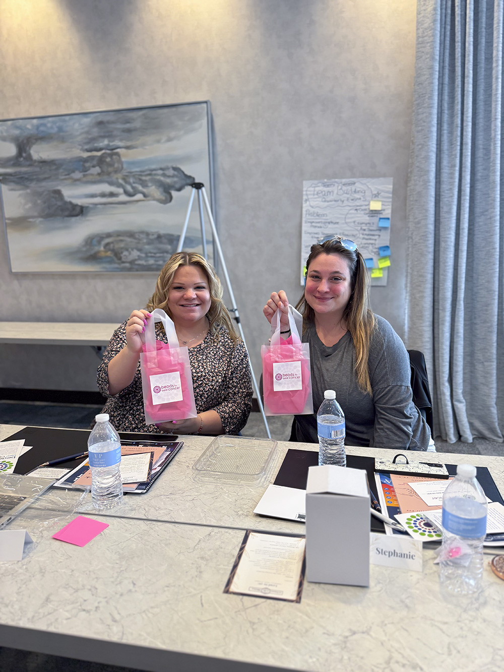 Two women holding up pink Beads to Beat Cancer gift bags at a Breast Cancer Awareness event