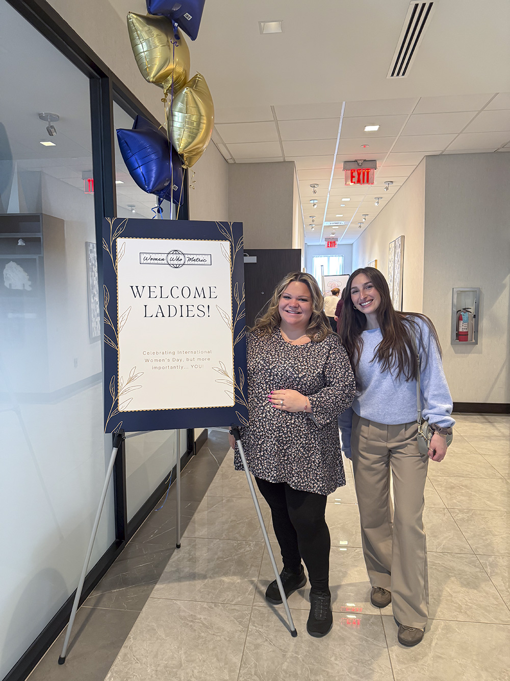 Two women posing next to a Women Who Metric Welcome Ladies sign with blue and gold balloons