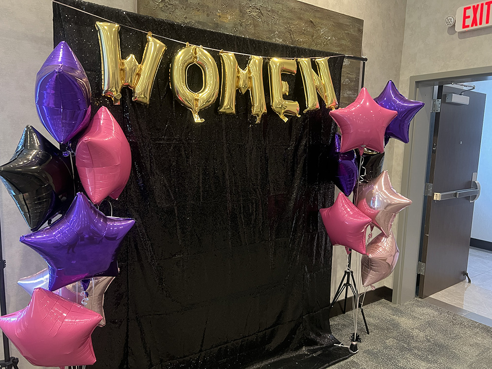 Gold WOMEN balloon letters above a sequin backdrop with pink and purple star balloons at a Women Who Metric event