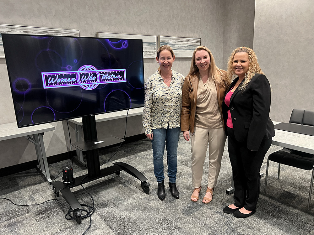 Three women posing next to a screen displaying the Women Who Metric logo